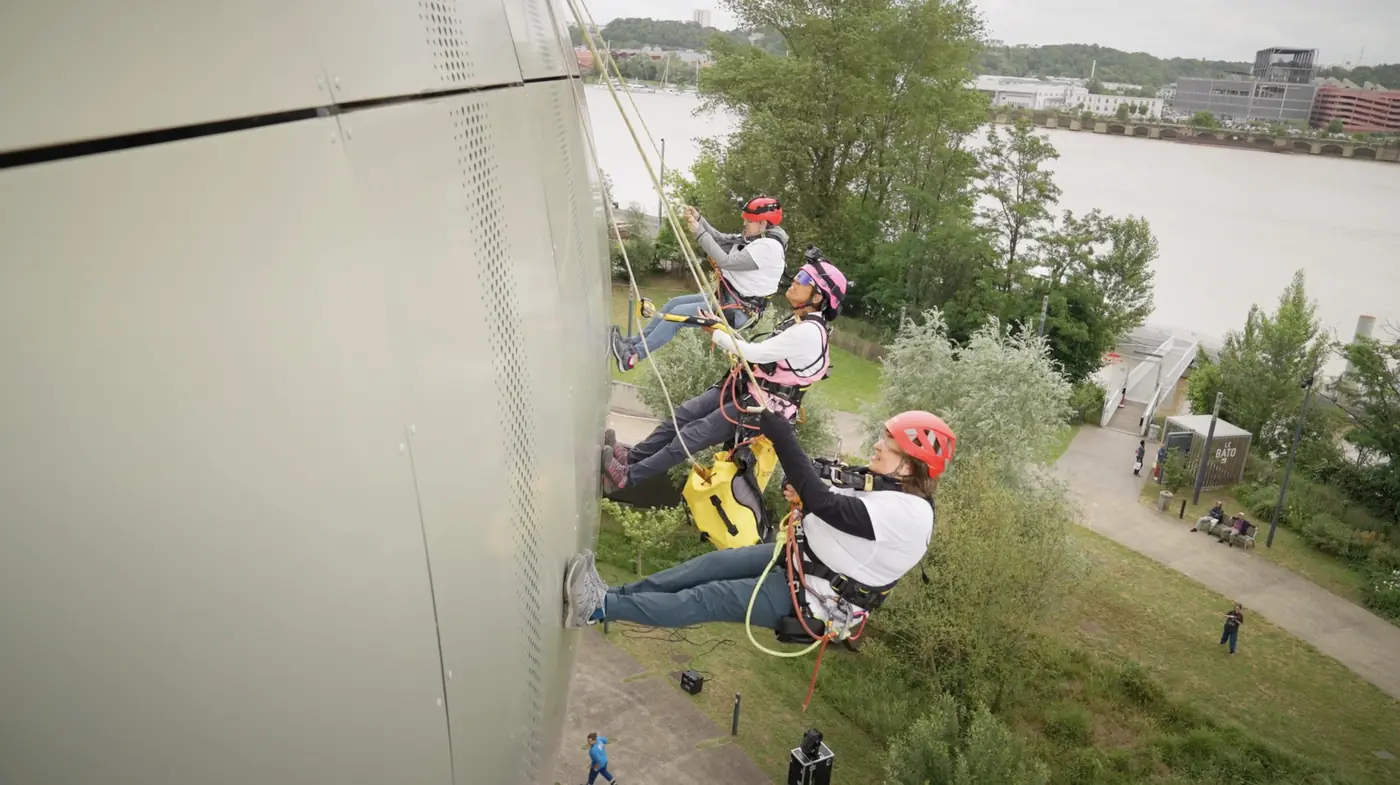 Trois Athl&egrave;tes du Vertige en rappel le long d'une fa&ccedil;ade m&eacute;tallique, casques rouges et rose, au bord d'un fleuve.